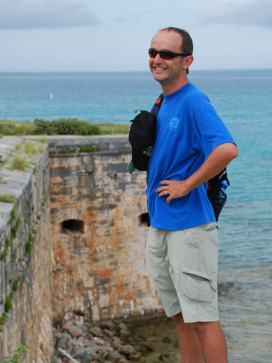 Photograph of Guy standing by the coast, smiling, wearing sunglasses and a blue T-shirt.